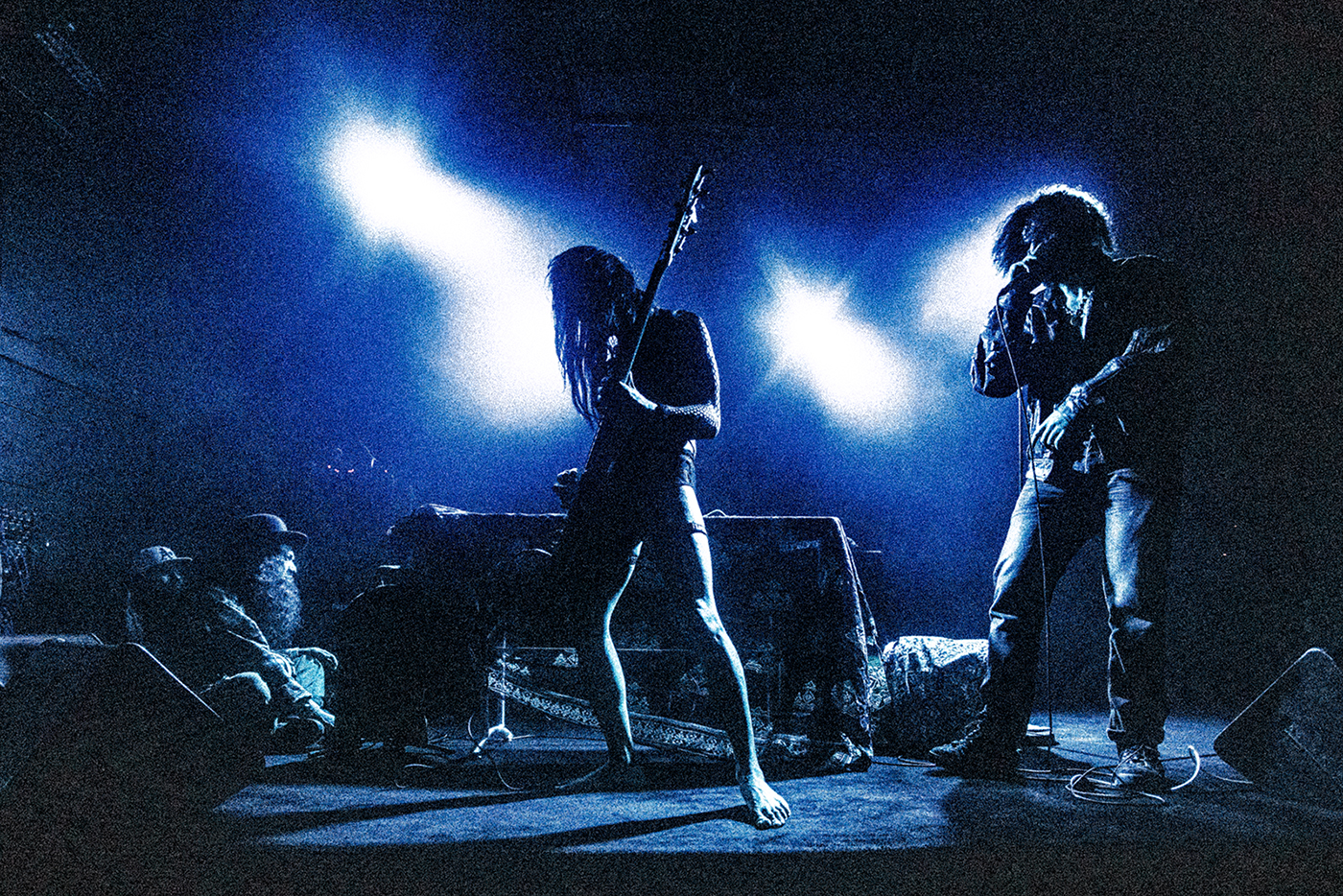 A dark, grainy photo of two performers on stage. One is playing bass and the other is singing.